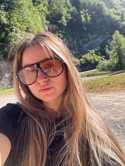 A young woman with long, blonde hair is posing for a selfie outdoors. She is wearing large, dark-rimmed sunglasses with orange lenses and a black t-shirt. The background shows a lush, green, forested hillside with a rocky cliff face and a road winding through it. The ground in the foreground is a gravelly dirt path. The lighting suggests it is a sunny day. There is no indication that this location is in L'Essard, France.