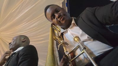 Two men in formal attire with a trombone. The man in the foreground is positioned at an angle, directing his gaze toward the viewer. He wears a white shirt with a black suit jacket, holding a trombone. The second man, situated behind and to the left, also sports a black suit jacket over a white shirt. A backdrop of creamy white fabric adds a soft, diffused light.