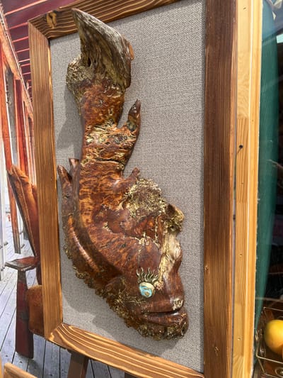 An artwork, possibly a carving or sculpture of a fish-like or humanoid head made from wood, is displayed in a frame against a textured gray background. The wooden artifact has a rough, organic texture with visible grain patterns and some golden accents. A small circular embellishment, resembling an eye with sun-like rays, is attached to the front of the artifact. The framed artwork is positioned outdoors, near a window, with a hint of a reflective surface and some yellow fruit in a basket visible in the background. The overall scene suggests an outdoor display or shop window in Yakunino, Russia.