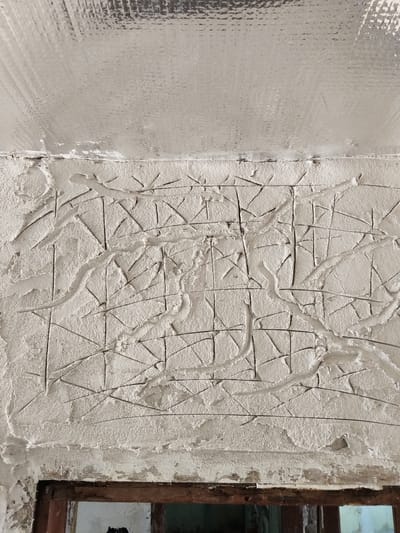 The image shows a wall with a textured finish, possibly plaster or stucco, with a pattern of incised lines. The lines are a mix of straight and curved, forming a grid-like structure with some irregular additions. The texture of the wall is rough and uneven, with some areas appearing more heavily applied than others. Above the textured wall, there is a reflective surface, possibly insulation or a metallic ceiling material, with a ribbed texture. The bottom of the image shows the top edge of a wooden structure, likely a doorframe or window frame, with some peeling paint and distressed wood. The overall impression is of a wall under construction or renovation. There is no text visible in the image. The lighting suggests an indoor setting, possibly during the daytime due to the brightness of the reflective surface. The context of Yavrovo, Bulgaria, provides a geographical location for the scene.