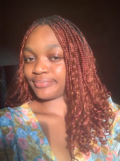 An image depicts a young adult female, appearing to be of African descent, positioned centrally and facing the viewer. Her skin tone is medium-dark, and she has a gentle, closed-lip smile. Her hair is styled in numerous reddish-brown box braids that extend below her shoulders, with the ends exhibiting a curly texture. She wears a garment featuring a floral pattern with hues of light blue, pink, yellow, and green, visible around her upper chest and shoulders. Strong, natural light illuminates the left side of her face and hair, creating highlights. The background is largely dark and out of focus, with a faint, indistinct pattern visible behind her left shoulder. The specific location within the city of Anantigha, Nigeria, is not discernible from the visual information presented.