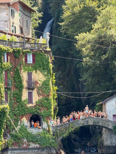The image depicts a sunny day in the village of Careno, Italy, overlooking the village's iconic waterfall and bridge. A weathered building, draped in vibrant green ivy, dominates the left side of the frame. Red-shuttered windows punctuate its facade, and an arched opening on the lower level reveals a group of people gathered inside. A balcony with a railing, adorned with statues and umbrellas, sits above this opening.
A stone bridge arches gracefully across a rocky gorge in the midground, its surface crowded with a large group of young men and women. Many are dressed in swimwear, suggesting they are preparing to jump into the water below. Some are looking out, while others are engaged in conversation.
Below the bridge, the rocky riverbed is visible, with a few individuals standing in the shallow water. In the background, a powerful waterfall cascades down a lush, tree-covered cliff face, adding a dramatic natural element to the scene. The overall impression is one of a lively summer gathering in a picturesque, historic Italian setting.