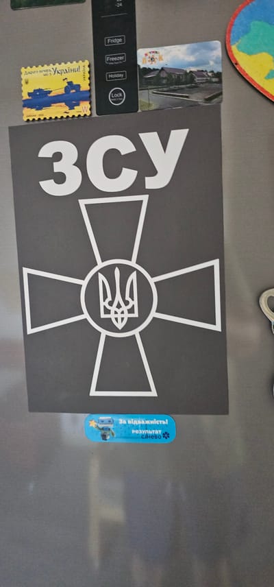 This is a photograph of a refrigerator door covered in various magnets and stickers. The central focus is a large, dark gray rectangular magnet featuring a white outline of a Maltese cross with the Ukrainian trident symbol in the center. To the left of this, a smaller yellow magnet in the shape of a postage stamp displays the Ukrainian flag colors and text in Ukrainian that translates to "Good evening, Ukraine!". Below this is a strip of buttons labeled "Fridge", "Freezer", "Holiday", and "Lock". To the right of the main cross magnet is a thin, blue vertical sticker with Ukrainian text that reads "For courage! The result is Sinevo." and depicts a cartoon astronaut. A small, rectangular magnet with a photograph of buildings and a blue sky is visible to the upper left of the main magnet. The overall impression is of a personalized refrigerator door, likely in a home setting in Berestyn, Ukraine, reflecting Ukrainian national pride and possibly a sense of resilience. The lighting suggests an indoor environment, possibly during the daytime. There are no people visible in the image.