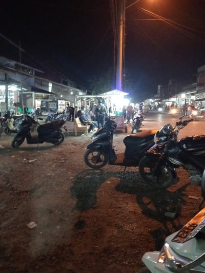 The image shows a night scene in Kruengmanyang, Indonesia. The location appears to be a roadside area with small shops or stalls lining the street. Several motorcycles are parked on a dirt or gravel surface. Some people are sitting on benches near one of the stalls, while others are standing or walking around. A tall utility pole is visible. Streetlights provide illumination, and the sky is dark.