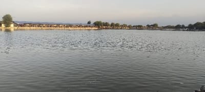 The image displays an expansive outdoor scene in Tambak Cemandi, Indonesia. A large body of water, likely a pond or *tambak*, occupies the majority of the frame, its surface exhibiting gentle ripples and reflections. Along the distant bank, a prolonged series of interconnected structures is visible. These structures are open-sided pavilions or shelters, characterized by their roofs and support posts, extending horizontally across the landscape. Within these pavilions, numerous individuals are present, appearing to be seated or congregated. On the far left of the bank, a light-colored building stands next to a single tree. Further to the right, additional trees are interspersed among the structures, and a wooden pier or walkway extends over or alongside the water in the distance. The sky above is light and hazy, consistent with either early morning or late afternoon illumination.