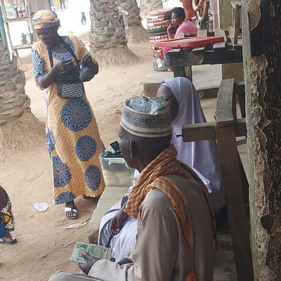 The image captures an outdoor scene, likely a market or street stall, under natural daylight. The ground is uneven, composed of dirt or sand. Several individuals are present, engaged in what appears to be a commercial or social interaction.
In the mid-ground, a man stands on the right side of the frame. He is wearing a light brown or beige traditional tunic, an orange and white checkered scarf draped over his shoulders, and a light-colored traditional cap (possibly a Fula cap or similar regional headwear) with blue and white patterns. He has a dark complexion and is looking downwards. In his right hand, he holds a stack of green banknotes, which appear to be Nigerian Naira denominations (possibly 500 or 1000 Naira notes due to their color and size).
To the left of this man, another woman stands, facing slightly towards the right. She is wearing a vibrant yellow dress adorned with circular blue and white patterns, a common motif in African wax print fabrics (Ankara). She also wears a matching headwrap. She is holding a small, dark-colored handbag, possibly made of woven material, in her left hand. Her feet, clad in simple white sandals, are visible on the dirt ground.
In the background, behind the standing man, a person is partially visible, seated or crouching. This individual is dressed in plain white attire and also appears to be holding green banknotes in their hand, mirroring the man in the mid-ground.
Further into the background, towards the left, another person, possibly a young girl or woman, is visible behind a wooden structure. She is wearing a pink top and appears to be tending to a stall. Various items, including what look like stacks of black tires or large circular objects, are arranged on a red surface in front of her.
The scene is framed by rough wooden structures, consisting of beams and posts, suggesting makeshift market stalls or shelters overhead and in the background. The overall atmosphere appears to be a typical daily activity in an outdoor setting. No specific text is clearly legible on any objects or surfaces.