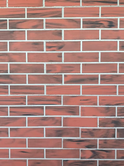 A close-up, eye-level shot captures a wall constructed of rectangular red bricks with dark, streaky markings. The bricks are laid in a staggered pattern, with thin, white grout lines separating them both horizontally and vertically. The wall appears to be an exterior surface, possibly part of a building. The lighting is even, suggesting it could be daytime, and there are no indications of specific weather conditions. No people or text are visible in the image. The setting is implied to be an outdoor environment, given the nature of the brickwork. The image provides no specific clues about the time of day, time of year, or geographical location beyond what is provided by the subject matter itself, which is a generic brick wall.