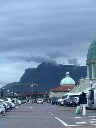 An overcast day in Milnerton, South Africa, features a mountain backdrop partially obscured by clouds. A street lined with white vans is seen along the right side, with the rear of the vehicles facing toward the viewer. A person wearing a dark jacket and light-colored pants walks on the sidewalk, carrying a green and white bag. Buildings with green and white domes line the street, appearing behind and adjacent to the vans. A yellow construction crane is visible against the mountain. The street is marked with white lines and what appear to be parking instructions.