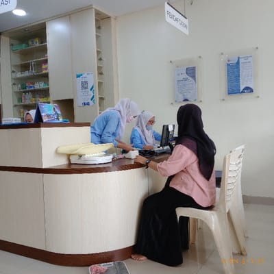 The image shows a scene inside a clinic or medical office in Kecamatan Curug, Indonesia (latitude: -6.263, longitude: 106.558). A reception desk with a white laminate countertop and wood trim is in the center of the image. Four women are present. Three women in headscarves are interacting with the counter. A woman wearing a pink shirt and a black skirt, holding a phone in her right hand, stands at the reception desk. A second woman wearing a blue headscarf and a light blue shirt is seated on a white chair, speaking with the woman in pink. A third woman wearing a light green headscarf and a white shirt, is seated in the background, interacting with a computer screen. A white scale and a baby scale are on the reception desk. A shelf with medicine bottles and a sign with a QR code are on the wall to the left. The wall to the right displays a sign in Indonesian that says "Registration" along with two informational posters. There is a plastic chair with a white frame in the image. The office is well lit.