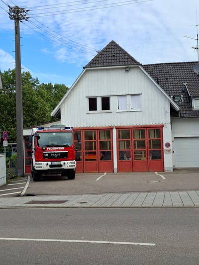 The image captures a fire station in Sinzheim, Germany. A red fire truck, identified by "FEUERWEHR" on its front, is parked in front of the station's entrance. The station building is white with a dark tiled roof and features several large red doors with windows. To the left of the station, a tall utility pole supports multiple overhead wires. In the background, lush green trees frame the scene, and a bright blue sky with scattered clouds is visible. The foreground shows a paved area and a road. A traffic sign with two red arrows is visible near the utility pole.
