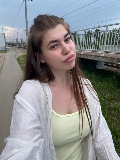 A young Caucasian woman with long, dark brown hair pulled back at the crown of her head is pictured from the chest up. She is wearing a light yellow tank top under a sheer white open-front shirt. The woman is standing on a concrete walkway next to a set of railroad tracks. A raised train platform with a white railing is visible to her right, with trees in the background. Overhead power lines and poles are present, suggesting an industrial or transportation area. The sky is overcast. The woman's face is turned towards the camera with a slight tilt of her head and a hint of a smile. She has light blue eyes and is wearing light makeup. Her skin appears fair with some freckles. The overall setting seems to be a train station or a railway yard in a city, potentially Rechytsa, Russia, given the context provided.