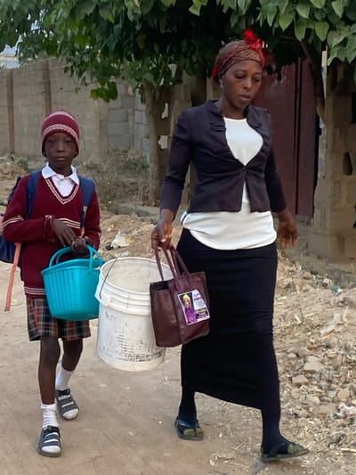 A woman in a black skirt, white top, and black jacket holds a brown purse while standing next to a white bucket. A child in a red sweater, plaid shorts, and white socks carries a blue basket. Both wear sandals. The background includes greenery, a building with a dark door, and a dirt or gravel surface. The image appears to be taken outdoors, possibly in a residential area.