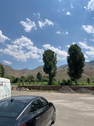 The image depicts a daytime scene in the city of Arpi, Armenia, with a focus on a landscape featuring mountains, vegetation, and vehicles. In the foreground, the rear section of a dark-colored sedan is visible, parked on a gravel lot. To the left, the side of a white van is present, displaying text that appears to be in Dutch, including "09 45 VAN DE MERBEL" and "Woninginrichting." Beyond the vehicles, a low stone wall runs horizontally across the frame, separating the gravel area from a lush green field. Further in the background, several tall, slender trees stand in front of a backdrop of rolling, arid mountains under a bright blue sky dotted with white, fluffy clouds. The overall impression is of a rural or semi-rural environment during fair weather.