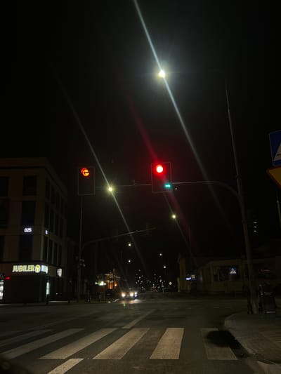 The image depicts a street scene at night in Siedlce, Poland. A prominent traffic light displays a red arrow indicating no left turn, and a green arrow indicating a right turn is permitted. To the left, a modern building with illuminated windows and the sign "JUBLIER NIKIES" is visible. Across the street, there are other buildings with lit windows and signage, suggesting a commercial area. A pedestrian crossing is clearly marked on the asphalt in the foreground. Streetlights and vehicle headlights illuminate the scene, creating streaks of light due to the low-light conditions and camera exposure. Several cars are present on the road, with headlights on. The overall atmosphere is that of a quiet urban environment at night, with functional street infrastructure and ongoing vehicle traffic.
