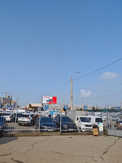 This image depicts an outdoor car market or large parking facility located in Eshanguzar, Uzbekistan, under a clear blue sky with minimal cloud cover. The foreground consists of cracked asphalt pavement and a light-colored metal fence, partially open on the right, where a mobile vendor stand with a metal barrel is positioned. The midground is occupied by a dense arrangement of vehicles, including white SUVs, dark sedans, and white commercial vans. Several individuals are present among the vehicles; one person is observed standing elevated on what appears to be the roof of a white van in the center-right. A light-colored streetlamp post with overhead power lines extends across the frame. In the background, a red and white billboard displaying "Kukmara" is visible above a multi-story white building labeled "ARUS XXIY CENTER." To the far left, an active construction site featuring a crane and partially completed structures is present, with additional buildings visible along the distant horizon.