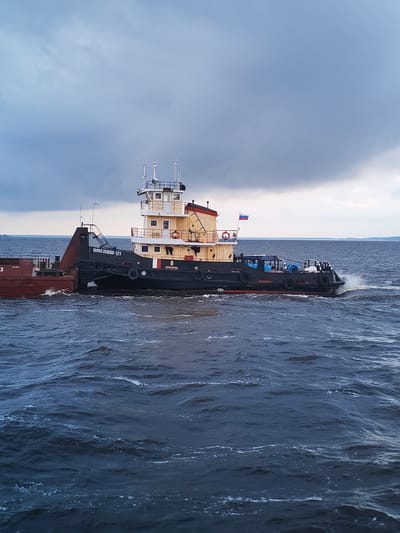 A tugboat named "ШКИПЕРОВ-121" is seen from a side angle, pushing a barge on a body of water under a cloudy sky. The tugboat is dark with a beige upper structure and a red stripe along its side. It has several windows and a Russian flag flying from its mast. The barge is reddish-brown. The water is choppy with whitecaps, indicating movement. The scene appears to be a waterway in or near Vichelovo, Russia, as suggested by the boat's name. There are no people visible in the image.