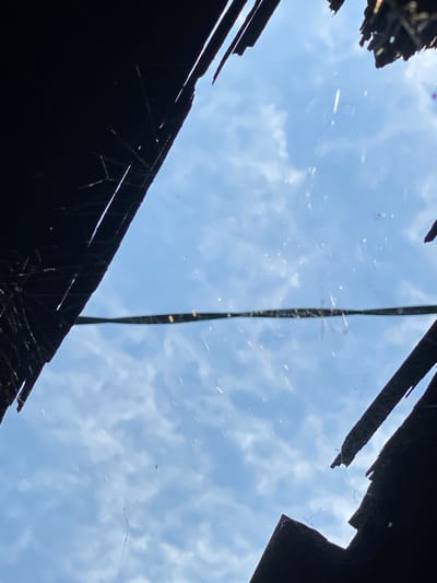 The image shows a close-up, upward-looking view through a broken roof or structure, revealing a bright blue sky with scattered white clouds. Dark, jagged silhouettes of the broken roof edges frame the sky, with thin strands of what appears to be spiderwebs visible across the opening. A horizontal, dark line, possibly a wire or debris, is stretched across the middle of the frame. The overall impression is one of decay and exposure to the elements. The location in Banjarmasin, Indonesia, suggests a tropical climate, which might explain the presence of spiderwebs and the bright sky. The scene depicts a damaged structure, possibly a dwelling or shed, with its roof partially collapsed, allowing a view of the sky. No people or specific actions are depicted.