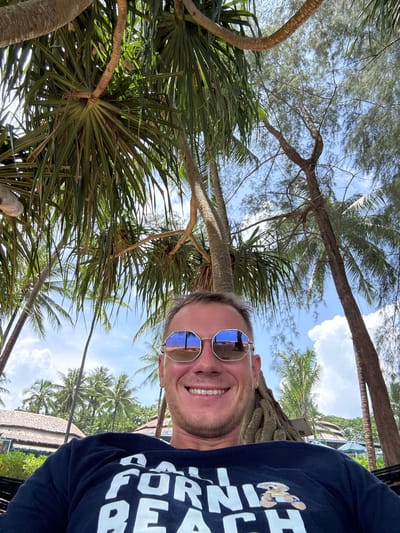 In Ban Khuk Khak, Thailand, a man in his 30s is lying in a hammock, looking at the camera. He is wearing mirrored sunglasses and a dark blue t-shirt with white lettering that appears to say "FORNI" and "BEACH". A cartoon bear is depicted on his shirt. The man is smiling and his face is framed by palm trees and other foliage. In the background, there are thatched-roof buildings, umbrellas, and more palm trees, indicating a resort or beach setting. The sky is blue with scattered white clouds. The reflection in the man's sunglasses shows a palm tree and a bench on a sandy area, further confirming the tropical beach location.