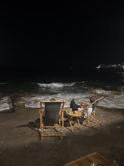 At night, two people sit in deck chairs facing the sea in Naousa, Greece. The waves crash against the shore, creating a foamy white spray. The figures are silhouetted against the dark water, with only faint outlines visible. One person is wearing a white striped shirt, while the other is dressed in dark clothing. A small wooden table sits between them, with drinks and ashtrays on it. In the distance, lights from buildings can be seen on the coast, suggesting the presence of a town or village. The scene evokes a sense of peace and relaxation, with the sounds of the ocean and the soft glow of the lights contributing to the tranquil atmosphere.