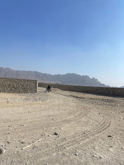 A desert landscape with tire tracks on the sandy ground. A low rock wall runs along the left side, with mountains fading into the hazy blue sky in the background. A person stands near a break in the wall. A single twig lies on the ground near the bottom right. The scene is bathed in bright sunlight.