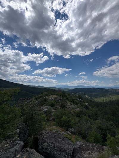 The image depicts a panoramic view from a rocky, elevated vantage point overlooking a vast, forested valley. The foreground is dominated by large, moss-covered rocks and sparse vegetation. The midground features a dense, dark green forest that covers rolling hills, transitioning into a hazy, layered landscape of more distant hills and mountains under a partly cloudy sky. The sky is a vibrant blue, punctuated by dramatic, white and grey cumulus clouds that cast shadows over the terrain below. The scene is serene and natural, suggesting a remote, mountainous location within the region of Dolno Drianovo, Bulgaria. There are no people or discernible man-made structures visible in the image.