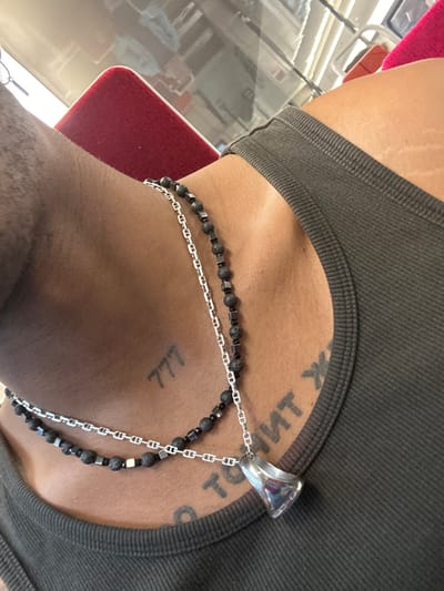 The image displays a close-up, angled view of an individual's upper torso, specifically the neck and upper chest, in Coye-la-Forêt, France. The subject has brown skin and is wearing a dark olive green ribbed tank top. Two layered necklaces are visible around the neck. The longer necklace consists of a silver-colored anchor-link chain strung with black porous beads and small square metallic beads. The shorter necklace, also a silver-colored anchor-link chain, features similar black porous beads and metallic beads, and suspends a polished, reflective, bell-shaped silver-colored pendant. A tattoo depicting the numbers "777" is visible on the left side of the neck/collarbone area. On the right side of the chest, beneath the clavicle, a partial black textual tattoo is present, appearing inverted or mirrored. The background is out of focus, revealing a bright red upholstered surface and reflections of an outdoor environment and interior structures (consistent with a window) in a glossy surface, indicating an interior vehicle setting.