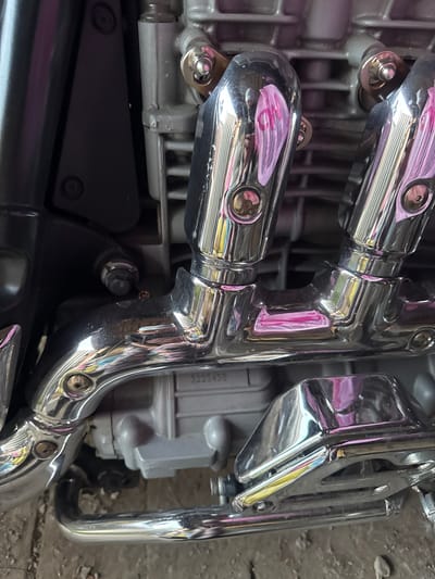 A close-up, eye-level, daylight, medium shot captures the chrome exhaust pipes of a motorcycle engine. The chrome reflects a pinkish hue and the surroundings, including what appears to be a blurry person in the reflection of the upper pipes. A grey engine block is visible behind the pipes, with a serial number that reads "3223458". The motorcycle is situated on a ground surface that appears to be concrete or asphalt, with a slight texture. The scene is set in Боголюбово, Russia.