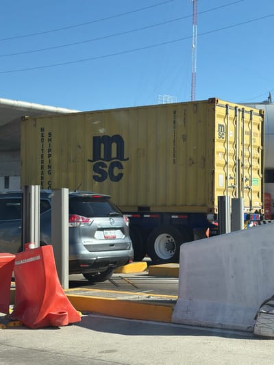 The image displays an outdoor scene in Santa María Moyotzingo, Mexico, under a clear blue sky with multiple power lines visible overhead. A large, yellow corrugated MSC shipping container is centrally positioned, marked with "MSC" logo, "MSMU 182565," "MSMU 18256," and "MEDITERRANEAN SHIPPING CO." This container is mounted on a dark blue truck trailer, exhibiting white-rimmed wheels. In front of the container, a silver Nissan X-Trail SUV, with a Mexican license plate reading "VMS-351-A," is partially obscured by two gray rectangular metal barrier posts. A second, darker-colored vehicle is partially visible to the left of the SUV. The immediate foreground contains two red plastic traffic barriers on paved ground, bordered by yellow painted curbs. A substantial light gray concrete barrier is positioned on the right, partially obscuring the truck trailer. In the background, a tall, red and white striped antenna tower is visible, along with a curved light-colored overhead structure on the upper left. No human individuals are present in the image.