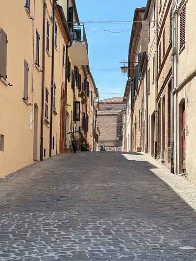The image depicts a narrow, upward-sloping cobblestone street in Barbara, Italy, lined with buildings on both sides. The buildings are constructed of brick and plaster, with facades painted in muted tones of yellow, beige, and ochre. Numerous windows with dark wooden shutters are visible. On the left side of the street, a building with a peach-colored facade features several windows and a small recessed doorway. On the right side, buildings display exposed brickwork and darker painted doorways. Hanging wires are strung across the street overhead. A bicycle is parked near the right-hand wall midway up the incline, next to potted plants. Further up the street, a red stop sign is visible on the right side. The sky is clear and blue. The sunlight casts strong shadows across the cobblestones, indicating a sunny day. The overall impression is one of a quiet, historic European street scene.