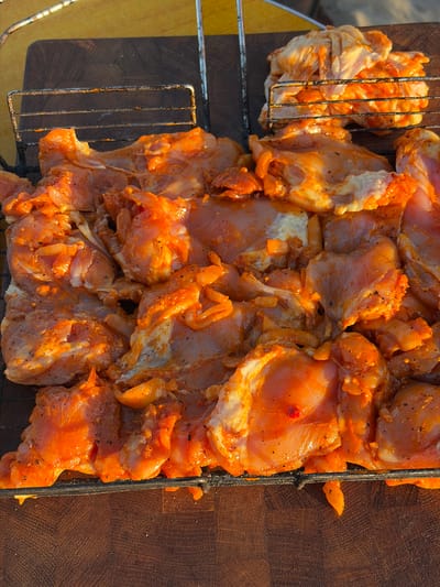 The image shows a close-up, overhead view of raw chicken pieces marinated in an orange sauce, arranged on a metal grill rack. The chicken is seasoned with black pepper. The grill rack is positioned on a dark wooden surface, possibly a cutting board or table. In the background, another grill rack with more marinated chicken is visible, stacked higher. The lighting suggests it is either early morning or late afternoon, casting a warm glow on the scene. The location is described as being in the city of Поваровка, Russia. There are no people or animals present in the image.