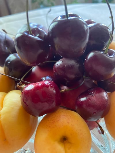 A close-up, eye-level shot features a clear glass bowl overflowing with ripe, dark red cherries and bright yellow apricots. The cherries, some with stems, are plump and glistening, with a few water droplets visible on their surfaces. They are densely packed, creating a vibrant contrast with the softer, rounder apricots nestled among them. The background is blurred, suggesting an indoor setting, but the natural light highlights the textures and colors of the fruit. The location of Askarovo, Russia, is not discernible from the visual information provided in the image.