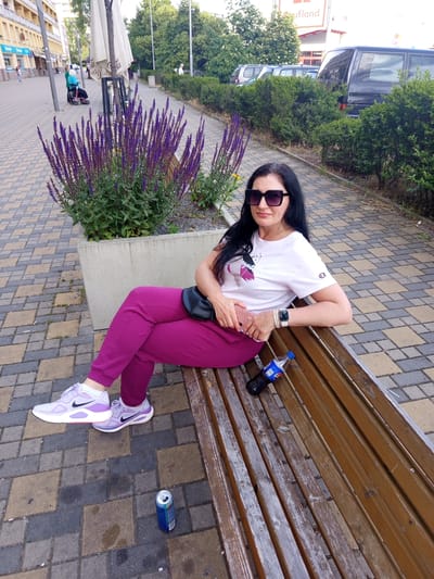 An adult female is seated on a weathered wooden slatted bench in an urban outdoor setting in Kladno, Czechia. She is positioned towards the right of the frame, looking directly at the viewer with a slight smile. Her hair is long, dark brown or black, and straight. She wears large, square-framed black sunglasses. Her attire consists of a white short-sleeved t-shirt with a purple and pink graphic design on the left chest and a red Champion logo on the left sleeve, magenta-colored joggers/sweatpants, and light purple athletic shoes with a black Nike swoosh. A black fanny pack is worn around her waist. On her left wrist, she wears a white-banded smartwatch and multiple light-colored bracelets. She holds a smartphone with a pink patterned case in her right hand.

To her right, a small plastic bottle of Pepsi with a blue cap rests on the bench. On the paved ground near her left foot is an empty blue and silver Monster energy drink can. Directly behind the bench is a large, rectangular concrete planter containing numerous tall, slender purple flowering plants, likely Salvia, along with green foliage and dark gravel.

The ground is paved with interlocking concrete pavers in shades of light brown, dark brown, and grey. To the left of the woman, a pedestrian sidewalk continues, bordered by multi-story light-colored buildings with windows and balconies. An outdoor café umbrella with a central pole is visible. Further down the sidewalk, a person in a blue top is seated in a red mobility walker/wheelchair, attended by another individual. Another person walks in the distance.

To the right of the woman, a row of green hedges separates the pedestrian area from a parking lot or street. Several vehicles, including a dark-colored van, are parked behind the hedges. Above the hedges and trees in the distance, part of a white sign with red lettering spelling "Kaufland" is visible. Numerous mature green trees are present in the background. The scene is illuminated by natural daylight, suggesting an overcast or late afternoon condition.