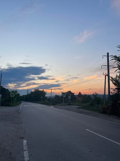 The image depicts a rural road in Solnechnaya Polyana, Russia, during twilight. The sky is a gradient of soft blues and oranges, transitioning from a clear expanse at the top to hues of pink and yellow near the horizon, indicating sunset. Scattered clouds, some dark and dramatic, others wispy and light, traverse the sky. The road itself is paved and appears to be a two-lane thoroughfare, curving slightly to the left as it recedes into the distance. White lane markings are visible on the asphalt.
On the left side of the road, a lamppost stands tall, with a street light illuminated. Adjacent to it, a pedestrian crossing sign with a yellow diamond shape and a white silhouette of a person is posted. Dense green foliage lines the left side of the road, with trees and bushes contributing to the natural landscape.
On the right side of the road, power lines are strung between tall utility poles. One pole is particularly prominent, with insulators supporting multiple wires. Further down the road on the right, a red-roofed building with a conical tower is visible amidst the trees. A piece of heavy machinery, possibly a loader with a yellow bucket, is parked on the right shoulder of the road, partially obscuring the dense greenery. The overall environment suggests a quiet, possibly developing, area on the outskirts of a settlement.