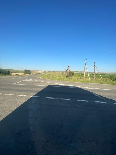 The image depicts a rural intersection under a clear blue sky. The foreground shows a paved road with white dashed lines and a triangular road marking. A dark shadow, likely from a vehicle, covers a portion of the road. In the middle ground, power lines stretch across the landscape, supported by wooden utility poles. The surrounding environment consists of grassy fields, some with hay bales, and scattered trees. Distant hills form the horizon. The location is identified as Moskovskoe, Russia.