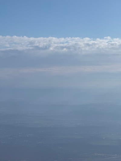 An aerial perspective from high altitude depicts a vast landscape under a partly cloudy sky, characterized by significant atmospheric haze. The upper third of the image shows a light blue sky. Immediately below this, a distinct horizontal band of scattered white cumulus clouds extends across the frame. Below these clouds, a dense layer of atmospheric haze obscures much of the mid-ground and foreground. The lower two-thirds of the image reveals an indistinct terrestrial surface, appearing as a sprawling mix of urban and suburban development interspersed with darker, possibly vegetated, areas. Due to the elevated viewpoint and atmospheric conditions, specific landmarks or detailed features identifying the city of La Garenne-Colombes, France, are not visually discernible. No individuals, identifiable objects, or specific actions are visible within the frame.