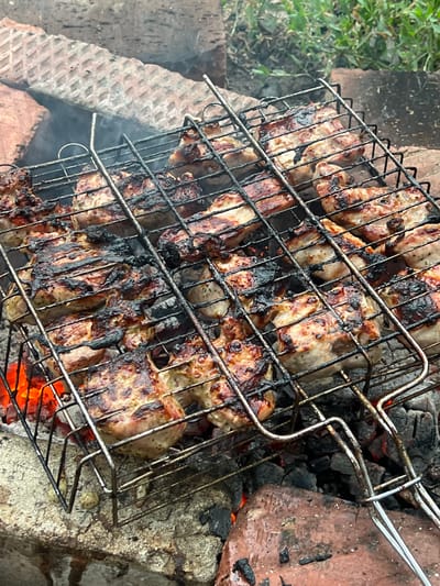 This image depicts a close-up, overhead view of meat cooking on a metal grill basket over hot coals. The meat appears to be chicken or pork, with some pieces showing charring and browning from the grill. Smoke is visible rising from the meat and coals. The grill is situated within a makeshift fire pit constructed from bricks and rocks. The background shows more bricks and some blurred green foliage, suggesting an outdoor setting. The scene is consistent with a barbecue or outdoor cooking event. There are no people visible in the image. The location in Kamen-na-Obi, Russia, is inferred from the provided context and is not visually discernible within the image itself.