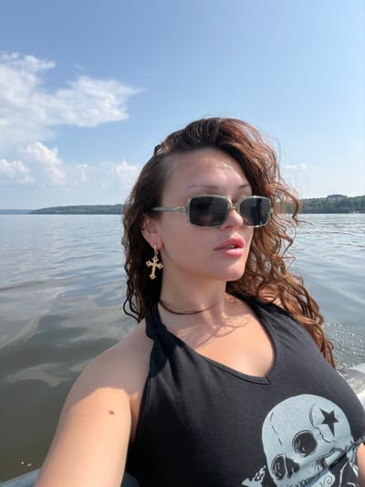 A woman wearing sunglasses and a black tank top with a skull graphic is on a boat. She has curly brown hair and is wearing a gold cross earring. The boat is on a body of water, with a tree-lined shore in the background. The sky is blue with white clouds. The location is the city of Sylva, Russia.