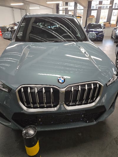 The image displays a stationary, muted gray-green BMW X-series SUV positioned centrally, facing the viewer, inside what appears to be a car dealership showroom or service area in Sprendlingen, Germany. The vehicle features a prominent dual kidney grille with vertical chrome slats and a BMW emblem on the hood. A black license plate holder is affixed to the lower front bumper, currently devoid of a license plate. The polished hood and windshield reflect overhead fluorescent lighting fixtures.
In the immediate foreground, a black and yellow striped bollard with a scuffed top is visible on the concrete floor, positioned slightly to the left of the vehicle's center.
The background reveals more of the interior environment. To the left, stacks of tires, an orange traffic cone, and other automotive equipment are partially visible, suggesting a workshop or storage area. To the right, large windows extend towards the ceiling, providing external light and a glimpse of what appears to be an urban building facade or street beyond. Parked further back in the showroom are at least two other vehicles: a blue SUV (likely another BMW) directly behind the featured vehicle, and a portion of a gray vehicle on the far right. The floor throughout the visible area is a light-colored concrete or epoxy surface with some visible wear and markings. No individuals are present in the scene.