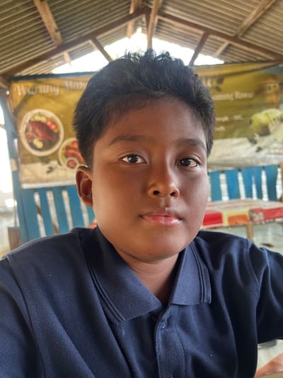 A front-facing portrait captures a child, male, with dark skin and short dark hair, positioned from the upper chest upward. The individual wears a dark blue collared polo shirt and looks directly at the camera. The background, out of focus, details an establishment identified as a 'warung' located in Gembong, Indonesia. A yellow banner with the text "Warung Maha" and images of prepared food, including a grilled fish dish, is partially visible. Structural elements of the environment include a corrugated metal roof, exposed wooden rafters, and blue wooden slatted partitioning. A table with a multi-colored patterned cloth (red, orange, white, yellow sections) is also present. The scene is illuminated by daylight.