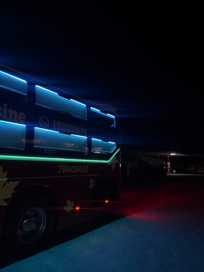 This image depicts a dark, outdoor scene, likely at night, with a bus prominently featured. The bus is dark blue with illuminated sections along its side, glowing with a bright blue light, possibly from LED strips. These illuminated sections appear to be windows or decorative panels. To the right of these panels, there is a section of the bus with yellow text. Some of the text is obscured, but visible words and numbers include "19002679" and "79H-094.52". There are also yellow leaf-shaped decals on the bus, suggesting a possible theme or branding.
In the background, a large white structure, possibly a building or a vehicle like a trailer, is visible, with some red lights reflecting on a surface, indicating another light source in the vicinity. The ground appears to be asphalt or concrete. The overall atmosphere is dim, with the strong blue lights of the bus being the most striking feature. There are no people visible in the frame. The red lights and the darkness suggest it might be late at night. The location context provided places this scene in M'Drắk, Vietnam.