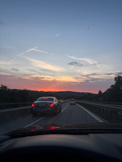 The image depicts a highway scene at dusk, likely in Hrusice, Czechia, as indicated by context. The perspective is from inside a vehicle, looking forward through the windshield. The sky is a gradient of blues and oranges, with contrails from aircraft forming an 'X' shape and other wispy clouds.
On the highway, several cars are visible. The most prominent is a dark-colored Mercedes-Benz sedan in the right lane, its brake lights illuminated. It displays a license plate reading "7AH 7888". Further ahead, other vehicles, including a silver SUV, are moving in the same direction. The highway is bordered by a metal guardrail on the right and dark trees and rolling hills in the distance. The road surface is asphalt, with yellow and white lane markings. The interior of the vehicle, including the dashboard and windshield, is partially visible in the foreground, reflecting the red glow of the brake lights. The overall atmosphere is that of evening traffic during a scenic sunset.