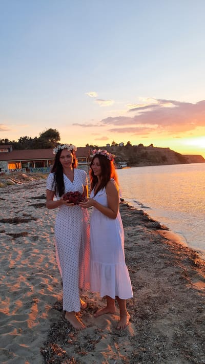 Two women stand on a sandy beach in Agia Triada, Greece, at sunset. The woman on the left wears a long white dress with black polka dots and a flower crown. She holds a shallow bowl filled with red berries. The woman on the right wears a white ruffled dress and a flower crown. Both women are barefoot. The sun is setting over the water, casting a warm orange glow across the scene. The sky is filled with colorful clouds. In the background, there are buildings and trees on a hillside. The waves gently lap at the shore.
