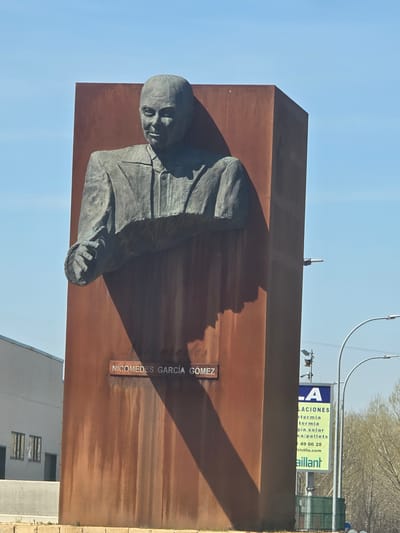 A large, rectangular monument, constructed from weathered, reddish-brown material, stands outdoors under a clear blue sky in Valverde del Majano, Spain. Embedded within the front face of the monument is a dark grey metal bust of a bald male figure, depicted from the chest up. The man has a slight smile and is wearing a collared shirt and jacket, with his right arm and partially formed hand extended forward. A prominent shadow cast by the bust extends diagonally downwards across the monument's surface. Below the bust, the name "NICOMEDES GARCÍA GOMEZ" is inscribed in white capital letters. To the left of the monument, a light-colored building with two barred windows is visible behind a grey wall. To the right, a street light with a curved arm is present, along with a partially obscured yellow-green billboard displaying text including "Vaillant". Bare trees and bushes are visible in the background further to the right.