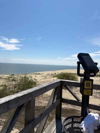The image depicts a scenic view of the Baltic Sea and sand dunes from a wooden observation deck. In the foreground, a coin-operated viewer is mounted on a black pole, with a white shirt draped over a nearby metal grate. The observation deck is made of weathered wooden planks and railings. Beyond the deck, vast sand dunes stretch towards the horizon, dotted with sparse vegetation. The sea is calm and blue, meeting the sandy shore under a clear sky with scattered white clouds. The location is the Curonian Spit in Morskoye, Russia, known for its unique landscape of shifting sands and coastal scenery.