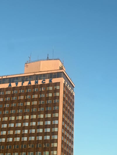 The image shows a tall, modern building with a repeating pattern of windows against a clear blue sky. The building has a beige facade and dark-framed windows, some of which appear to be open, creating a checkerboard effect. Blue lettering is visible vertically along one side of the building, spelling out "АЛТАЧСК" (ALTACSK in Cyrillic).

The building's structure suggests it is a hotel or an apartment complex. The angle of the shot, looking upwards from below, emphasizes its height. Antennas and other structures are visible on the roof, indicating it's a functional building. The bright sky suggests it is daytime, likely with clear weather. The overall scene is urban and industrial, with the building dominating the frame and the sky serving as a backdrop. There are no people visible in the image. The presence of Cyrillic text suggests the location is in a region where this script is used, consistent with Lobacheve, Ukraine.