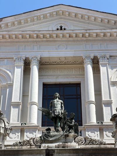 The image depicts the exterior of a building with classical architectural features, likely a church or public building, in Mozzo, Italy. The facade is made of white stone and features fluted columns with ornate capitals, a triangular pediment with decorative elements, and a large, dark-paned window. In front of the building, there is a bronze statue of a winged figure in military attire, standing over a possibly fallen figure or a base with drapery. To the left and right of the main statue, parts of other statues are visible, one on the left appearing to be a robed figure and one on the far right also in classical attire. The statues are positioned on a tiered stone base adorned with intricate carvings. The sky above is clear and blue, suggesting a sunny day. The overall impression is one of grandeur and historical significance.