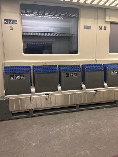 The image displays the interior of a train or public transport carriage, rotated 90 degrees clockwise, suggesting it's a dedicated area for luggage or bicycles. The setting appears to be an aisle or vestibule area, possibly at a station stop, given the concrete-like surface visible on the far right.

On the right side of the frame, a row of five foldable seats is visible, designed to maximize space when not in use. Each seat features a blue checkered fabric on the backrest and a gray seat cushion. Prominently displayed on each seat's backrest are three distinct symbols: a bicycle, a "no smoking" sign (a lit cigarette with a diagonal line through it), and a wheelchair user symbol, indicating that this area is designated for bicycle transport, is wheelchair accessible, and smoking is prohibited. Below the seats, a durable metal floor with ribbed sections and securing hooks can be seen, likely for anchoring luggage or bicycles.

To the left, a light-colored wall features a large rectangular window. Through the window, an interior view of another part of the train or an adjacent platform with more seating or luggage racks is visible. Reflections within the window show vertical striped patterns, possibly from blinds or another train's interior. Below the window, the same set of symbols (bicycle, no smoking, wheelchair access) is repeated, along with a QR code. Further up on the wall, near what appears to be a door or partition, a large "2" followed by a small human figure symbol is present, likely denoting a 2nd class compartment. A small rectangular sign with unreadable fine print is also affixed to the wall.

No people are present in the scene, and no specific activity is taking place. The lighting is bright and even, likely from interior lights, with a subtle glow through the window suggesting daytime. The overall impression is one of functionality and clear signage for passenger convenience and safety in a transport environment, consistent with trains operating in locations like Haßfurt, Germany.