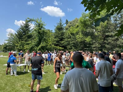 In the city of Razliv, Bulgaria, on a sunny day, a group of approximately fifty people are gathered on a well-maintained grassy field, bordered by mature trees and lush foliage. The gathering appears to be an informal outdoor event, possibly a competition or celebration. Several individuals are interacting around a white folding table, where a person in a blue shirt is handing a bottle to another person, suggesting a distribution or award ceremony. Others are observing or participating in activities. The attendees are dressed in casual summer attire, ranging from t-shirts and shorts to athletic wear. The atmosphere is lively and social, with people standing, talking, and some are in motion. A few vehicles are visible in the background, indicating proximity to a parking area. The sky above is a clear blue with scattered white clouds.