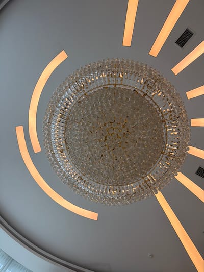 The image shows a close-up, high-angle view of a large, ornate crystal chandelier hanging from a white ceiling. The chandelier has a golden frame and is adorned with numerous clear, multifaceted crystals that reflect light. The ceiling features several linear, recessed lights emitting a warm, orange glow, arranged radially around the chandelier. One rectangular air vent is visible on the ceiling. The image does not provide any information that can be used to determine the location of the scene in Ankara, Turkey.