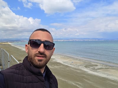 The image displays an adult male in a selfie perspective, occupying the left half of the frame. He has short dark hair, a beard, and is wearing dark sunglasses, a dark quilted vest, and a dark long-sleeved shirt. He is looking directly at the camera. The background depicts a coastal environment within the city of Unknown, Sovereign Base Areas of Akrotiri and Dhekelia. To the left, behind the individual, a metal barrier or temporary fencing is partially visible. A sandy beach recedes into the distance on the right, bordering a calm body of water. Across the water, a distant cityscape featuring multiple buildings, some appearing as high-rises, is visible against a backdrop of mountains on the horizon. The sky is partly cloudy, with areas of blue sky and scattered white and gray clouds.
